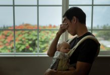 A depressed dad at home with his child covering his face and standing next to the window. Stay at home dad.