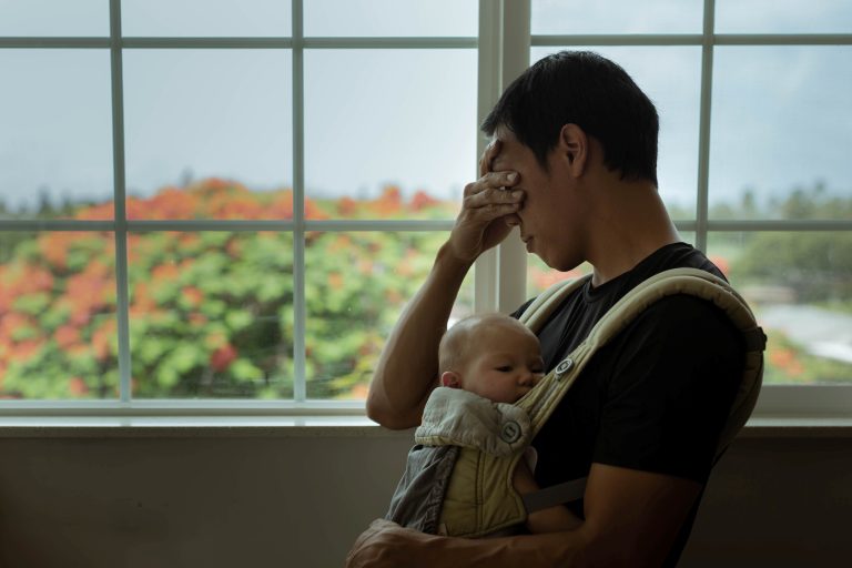 A depressed dad at home with his child covering his face and standing next to the window. Stay at home dad.